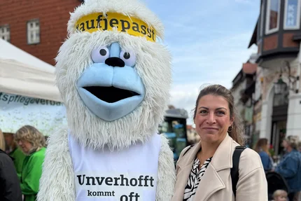 Das Kampagnenmasskottchen Rüdigga (Yeti) steht neben einer lächelnden Frau im beigen Mantel auf dem Umweltmarkt Wernigerode.