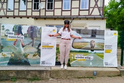Erxleben © MID Frau mit Fahrradhelm zeigt auf aufjepasst-Banner für Rücksicht auf Kinder und Abstand im Straßenverkehr.