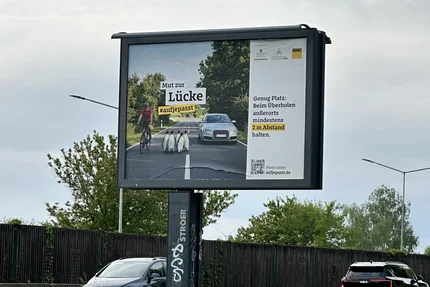 Großflächenplakat mit Radfahrer, Auto und Pinguinen, das den seitlichen Mindestabstand von zwei Metern beim Überholen außerorts anschaulich darstellt.