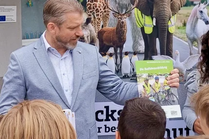 Calbe Saale © AGFK LSA Ein Mann zeigt Kindern das Heft „Die Radfahrausbildung“ vor einem Banner mit Verkehrssicherheitsmotiven.
