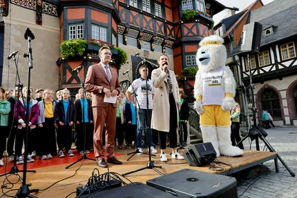 Bühne vor einem Fachwerkhaus in Wernigerode: Eine Frau spricht ins Mikrofon, daneben stehen ein Mann, ein Kinderchor und das Yeti-Maskottchen der Kampagne „#aufjepasst“.