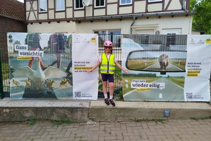 Erxleben © MID Kind mit Helm und Warnweste steht zwischen aufjepasst-Bannern zu Schulweg und Abstand.
