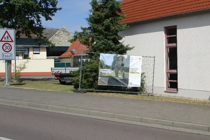 Wulfen © Gemeinde Osternienburger Land aufjepasst-Banner in Wulfen erinnert vor einem Gebäude an Vorsicht bei haltenden Bussen und Fahrgästen.